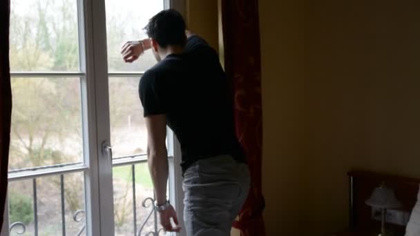 Young man standing against window, sad or worried, thinking — Stock Video