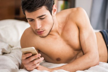 Yatakta cep telefonuyla yazan yakışıklı genç adam.