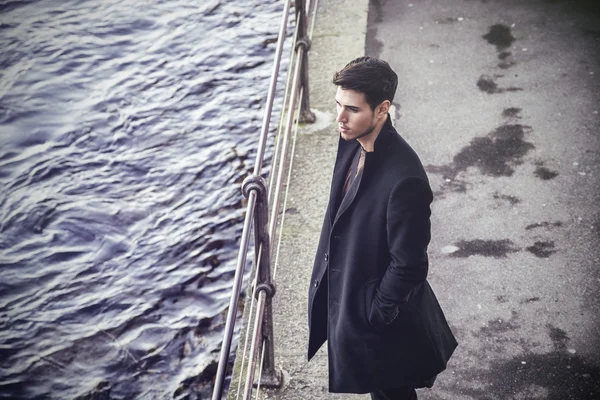 Young man in black coat looking down while leaning on railing - Stock-foto