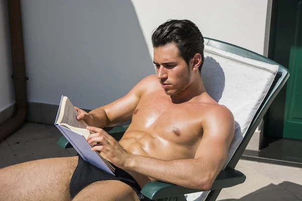 Genç adam Lounge sandalye okuma kitabı üzerinde güneşlenme — Stok fotoğraf