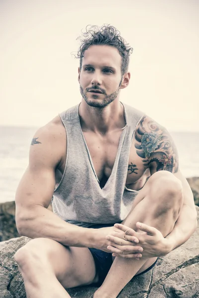 Hombre musculoso guapo en la playa sentado en las rocas —  Fotos de Stock