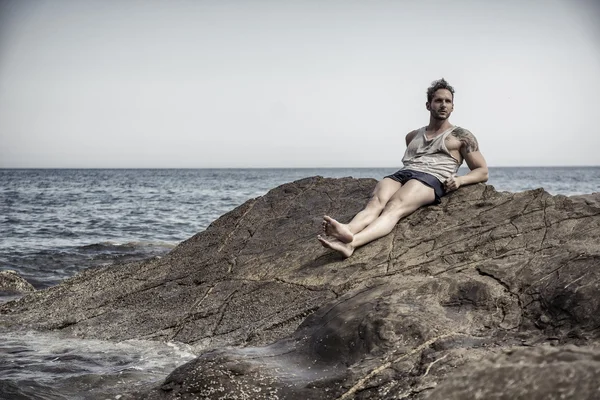 Bonito homem musculoso na praia sentado em rochas — Fotografia de Stock