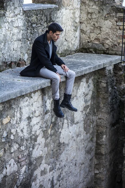 Retrato de hombre de pelo negro sentado en el borde de piedra y mirando hacia abajo —  Fotos de Stock