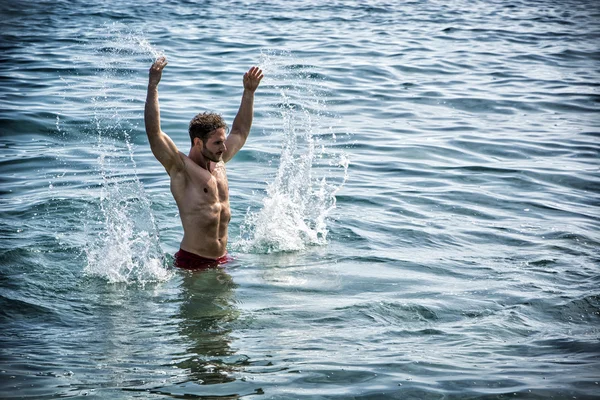 Handsome young bodybuilder in the sea, splashing water up — Stock Photo, Image