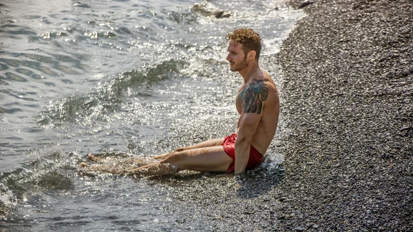 Hezký svalnatý muž na pláži ležící na štěrku — Stock fotografie