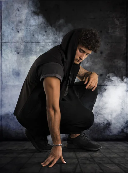 Handsome young man kneeling down in dark hoodie on black background — Stock Photo, Image