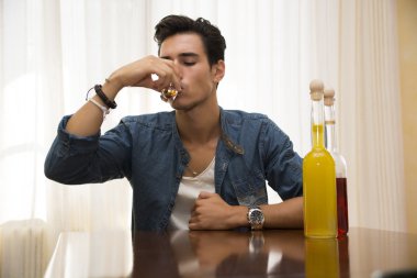 Young man sitting drinking alone at a table with two bottles of liquor clipart