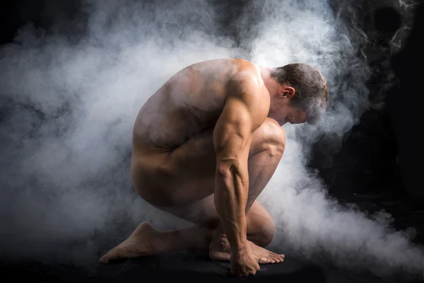 Schöner nackter Muskelmann kauert im Nebel im Studio — Stockfoto