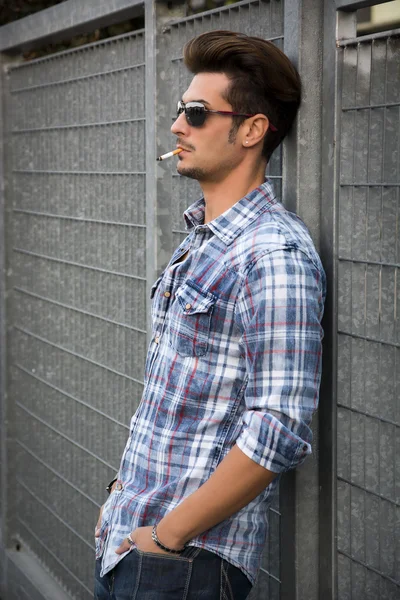 Trendy cool young man standing outside smoking, leaning against metal gate — Stock Photo, Image