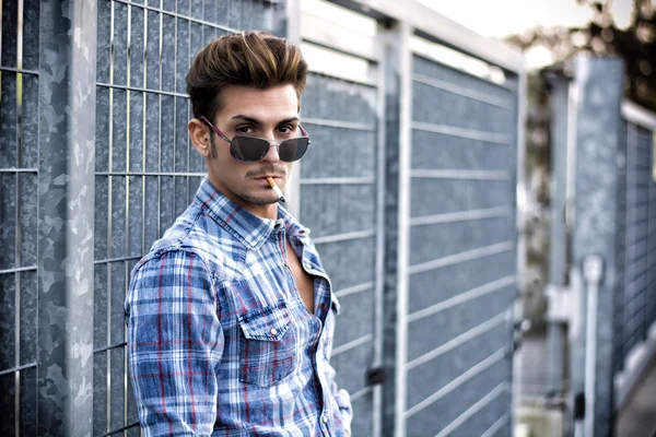 Trendy cool young man standing outside smoking, leaning against metal gate — Stok Foto