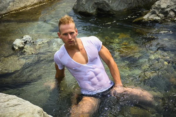 Giovane muscolare bello seduto nello stagno di acqua, indossando t-shirt bagnata — Foto Stock