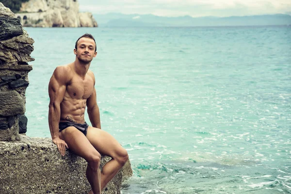 Giovane bello muscoloso sulla spiaggia seduto sulle rocce — Foto Stock