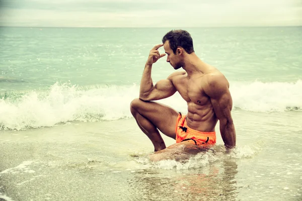 Handsome muscular young man on the beach sitting in the sea — Stock Photo, Image