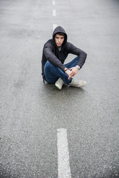 Νεαρός άνδρας στο μαύρο hoodie πουλόβερ κάθεται στη μέση του δρόμου — Φωτογραφία Αρχείου