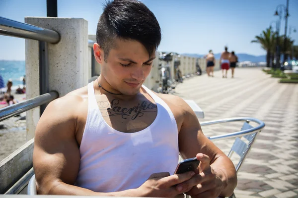 Όμορφος νεαρός άνδρας στο seaside κάθεται και χρησιμοποιώντας το κινητό τηλέφωνο — Φωτογραφία Αρχείου