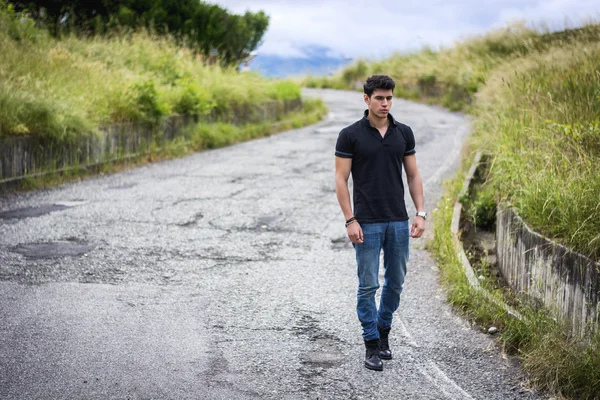 Young man in jeans and black t-shirt walking along rural road — Stock Photo, Image