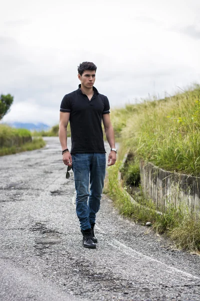 Hombre joven en vaqueros y camiseta negra caminando por camino rural —  Fotos de Stock