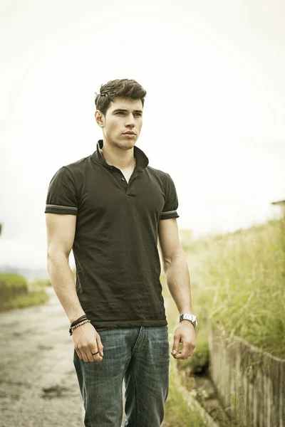 Young man in jeans and black t-shirt walking along rural road — Stock Photo, Image