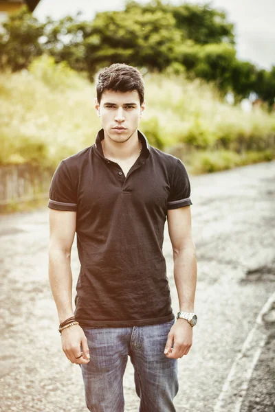 Young man in jeans and black t-shirt walking along rural road — Stock Photo, Image