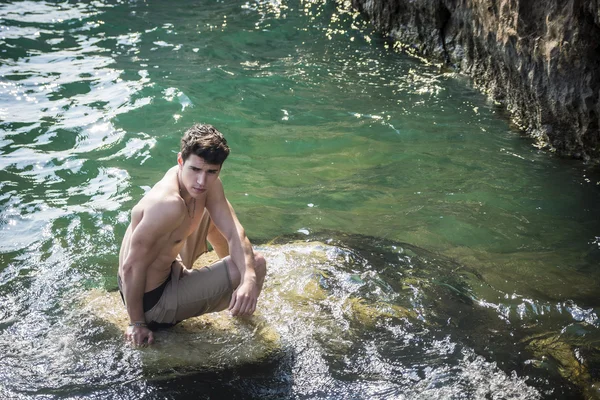 Young shirtless athletic man sitting on rock by sea — Stock Photo, Image
