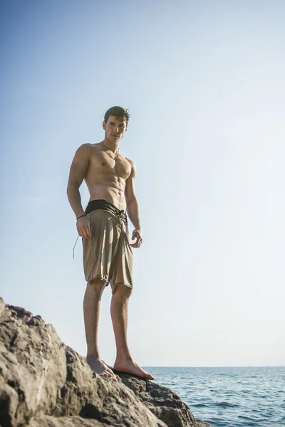 Young shirtless athletic man standing on rock by sea — Stock Photo, Image