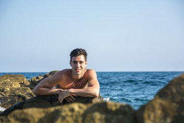 Young shirtless athletic man leaning on rock by sea