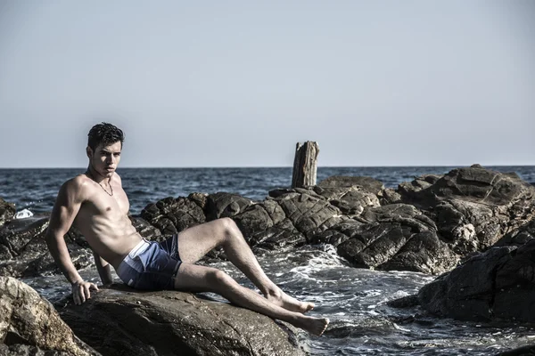 Young shirtless athletic man sitting on rock by sea — Stock Photo, Image