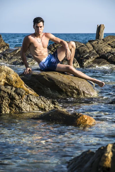 Young shirtless athletic man sitting on rock by sea — Stock Photo, Image