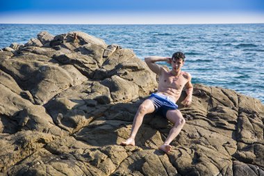 Young shirtless athletic man leaning on rock by water on ocean shore