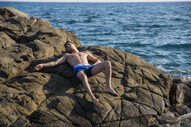 Young shirtless athletic man leaning on rock by water on ocean shore
