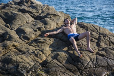 Young shirtless athletic man leaning on rock by water on ocean shore