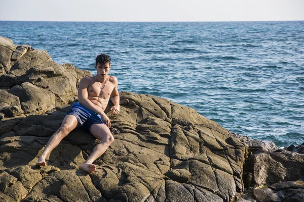 Young shirtless athletic man leaning on rock by water on ocean shore — Fotografia de Stock