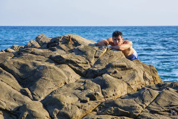 Young shirtless athletic man leaning on rock by water on ocean shore — Stock fotografie