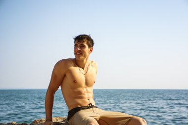 Smiling young man sitting on rock by sea or ocean shore