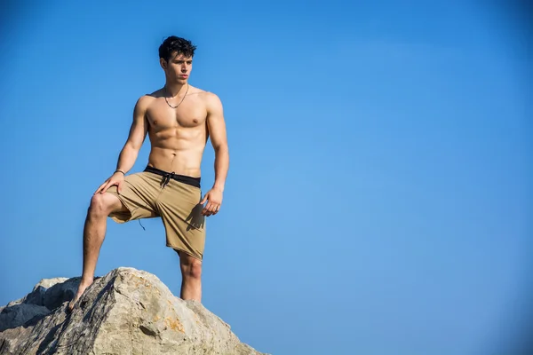 Young shirtless athletic man climbing on rock by water on ocean shore — Stok fotoğraf