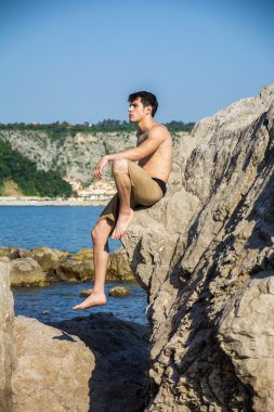Smiling young man sitting on rock by sea or ocean shore