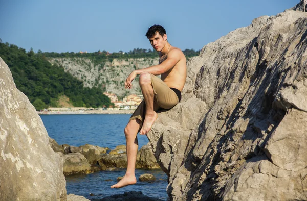 Smiling young man sitting on rock by sea or ocean shore — Stock Photo, Image