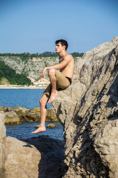 Smiling young man sitting on rock by sea or ocean shore — Stock fotografie