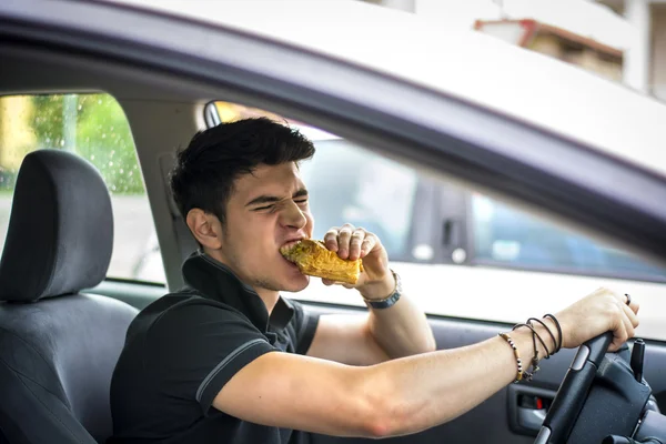 Unge man kör sin bil medan han äter mat — Stockfoto