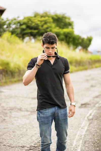 Young man in jeans and black t-shirt walking along rural road — Stock Photo, Image