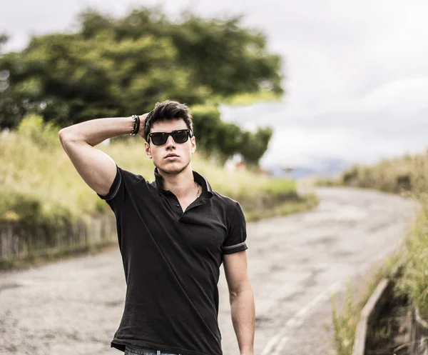 Young man in jeans and black t-shirt walking along rural road — Stock Photo, Image