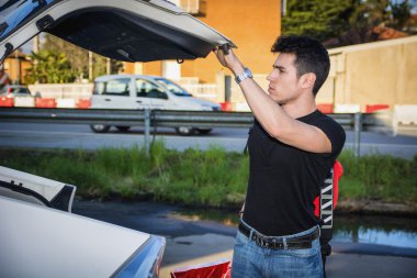 Young man taking luggage and bag out of car trunk