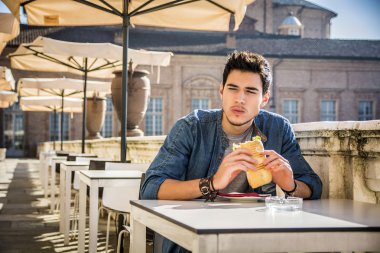 Genç adam kafede otururken sandviçini tutuyor.