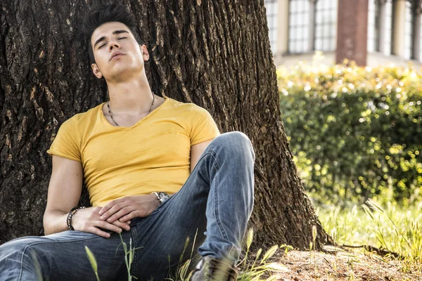 Aantrekkelijke jongeman in het park rustend tegen de boom — Stockfoto