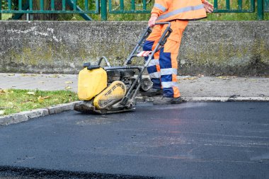 Paving işçisi kaldırım taşlarının yanındaki yeni asfaltı sıkıştırmak için titreşim plakası sıkıştırıcısı kullanıyor 