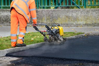 Paving işçisi kaldırım taşlarının yanındaki yeni asfaltı sıkıştırmak için titreşim plakası sıkıştırıcısı kullanıyor 
