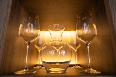 Şarap kadehleri ve cam sürahi, ahşap bir rafta simetrik bir şekilde sergileniyor. Kaliteli bir yemek salonunda, arka planda altın bir ışık var..