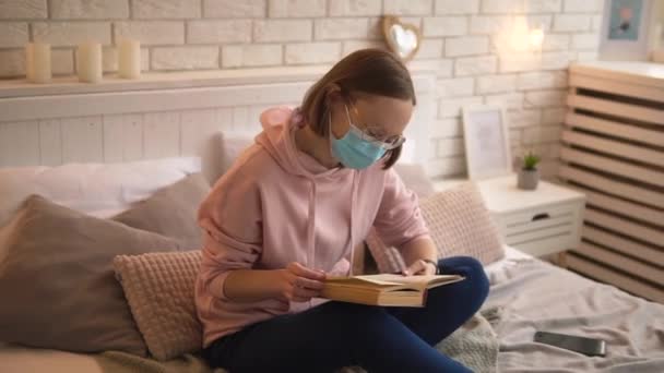 Случайная девушка в медицинской маске и в очках больная ковид-19 читает книгу на кровати дома. — стоковое видео