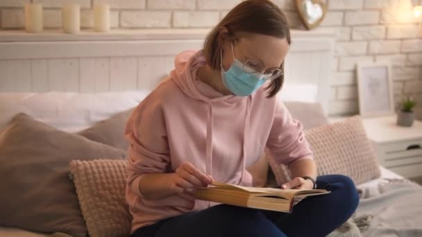 医療マスクの少女がベッドの上に座っている本を読む. — ストック動画
