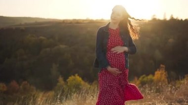 Genç hamile bir kız tepenin üstünde duruyor, saçları rüzgarda dalgalanıyor..